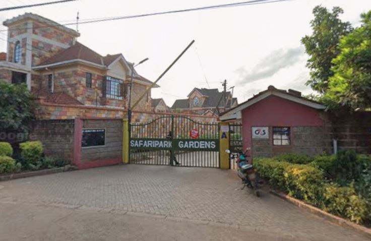 A gated entrance to Safaripark Gardens, with brick buildings and lush greenery. A person stands near the gate, and a motorcycle is parked nearby.