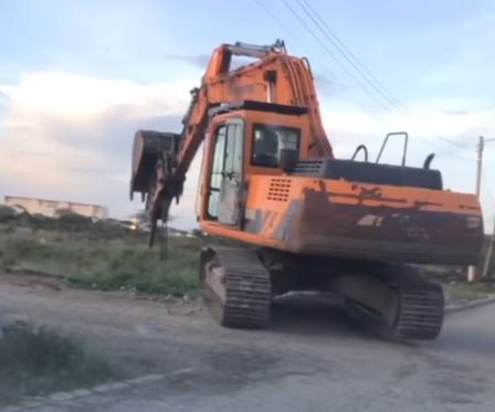 An orange excavator on a dirt road in a field, with a cloudy sky backdrop. It's stationary, no visible text or people present.