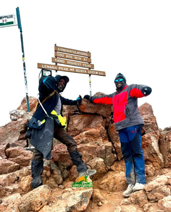Two climbers pose triumphantly at Lenana Peak, Mt. Kenya. Signs read "Congratulations" and details about the peak. Rocky summit, cloudy sky.