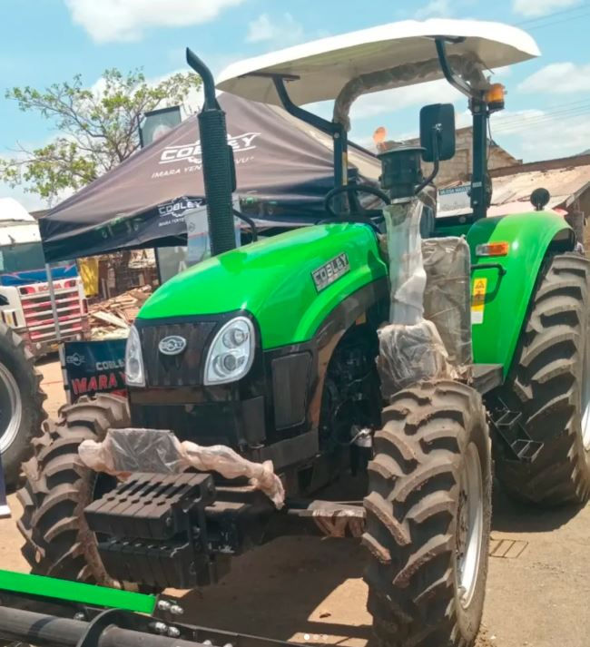 Bright green COBLEY tractor on display outside, wrapped in protective plastic. A black tent in the background shows the brand "COLEBY." Sunny day.