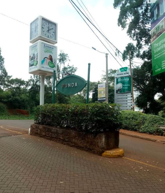 Runda sign with lush greenery on a roadside. A clock tower with ads, and several signposts visible against a cloudy sky. Calm atmosphere.