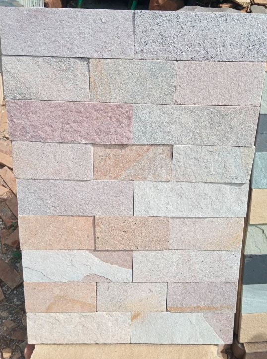 Stacked rectangular stone tiles in soft gray, beige, and pink tones displayed outdoors as a sample wall pattern