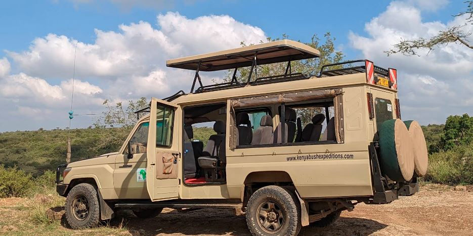 Beige safari vehicle with open roof and doors, parked in a grassy landscape at Nairobi National Park under a blue sky with clouds. Text: www.kenyabushexpeditions.com.
