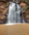 Ngare Ndare Waterfall cascading over rocky cliffs into a brown pool, surrounded by greenery. A tree branch frames the top. Serene, natural setting.