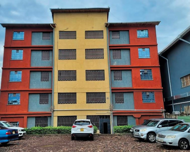 Nanyuki cozy haven   with yellow and red sections, windows barred. Parked cars in front on a gravel lot, cloudy sky above.