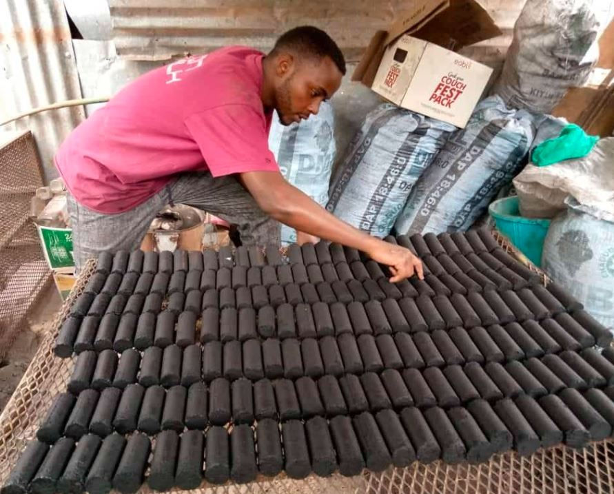 Man in pink shirt arranges black briquettes on a metal rack, surrounded by stacked bags and boxes. Industrial setting. Focused mood.