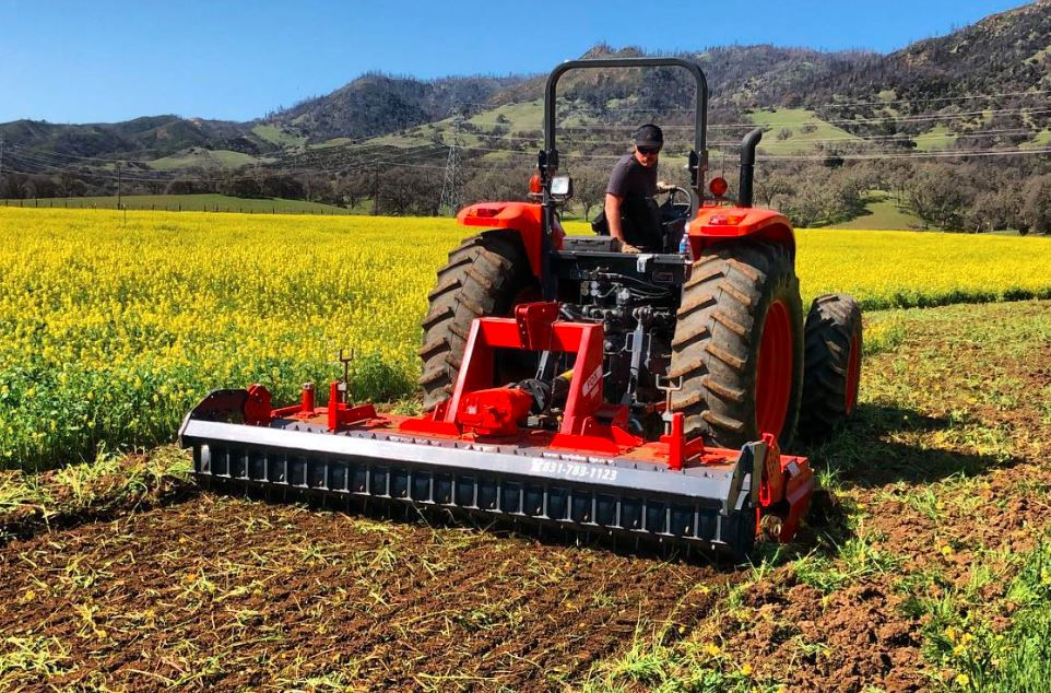A man drives a red tractor through a bright yellow field, plowing soil under a clear blue sky with mountains in the background.