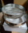 Two stainless steel Honey Strainer sieves stacked on a white box, set against a cardboard background. The setting is indoors, with a utilitarian vibe.