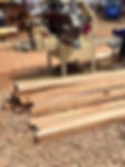 A sawmill machine cuts wood planks. Piles of fresh-cut timber lie on the ground. Workers operate equipment in a dusty outdoor setting.