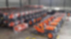 Orange kubota tractors lined up in a warehouse, some wrapped in plastic. A person inspects a tire. The mood is industrial and orderly.