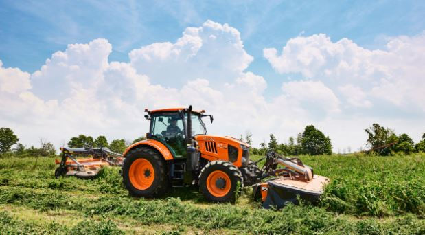 Orange tractor mowing grass under a clear blue sky with fluffy clouds. Green field and trees in the background, sunny and serene.