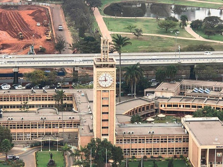 Aerial view of a large beige building with a clock tower, surrounded by greenery. Construction site and road in the background. Calm setting.