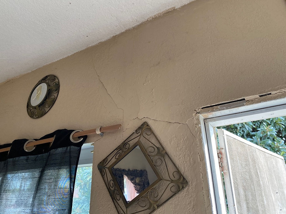 Beige wall corner with visible cracks, a decorative plate, a tilted mirror, and partially open window with dark curtains, suggesting disrepair. Foundation cracks.