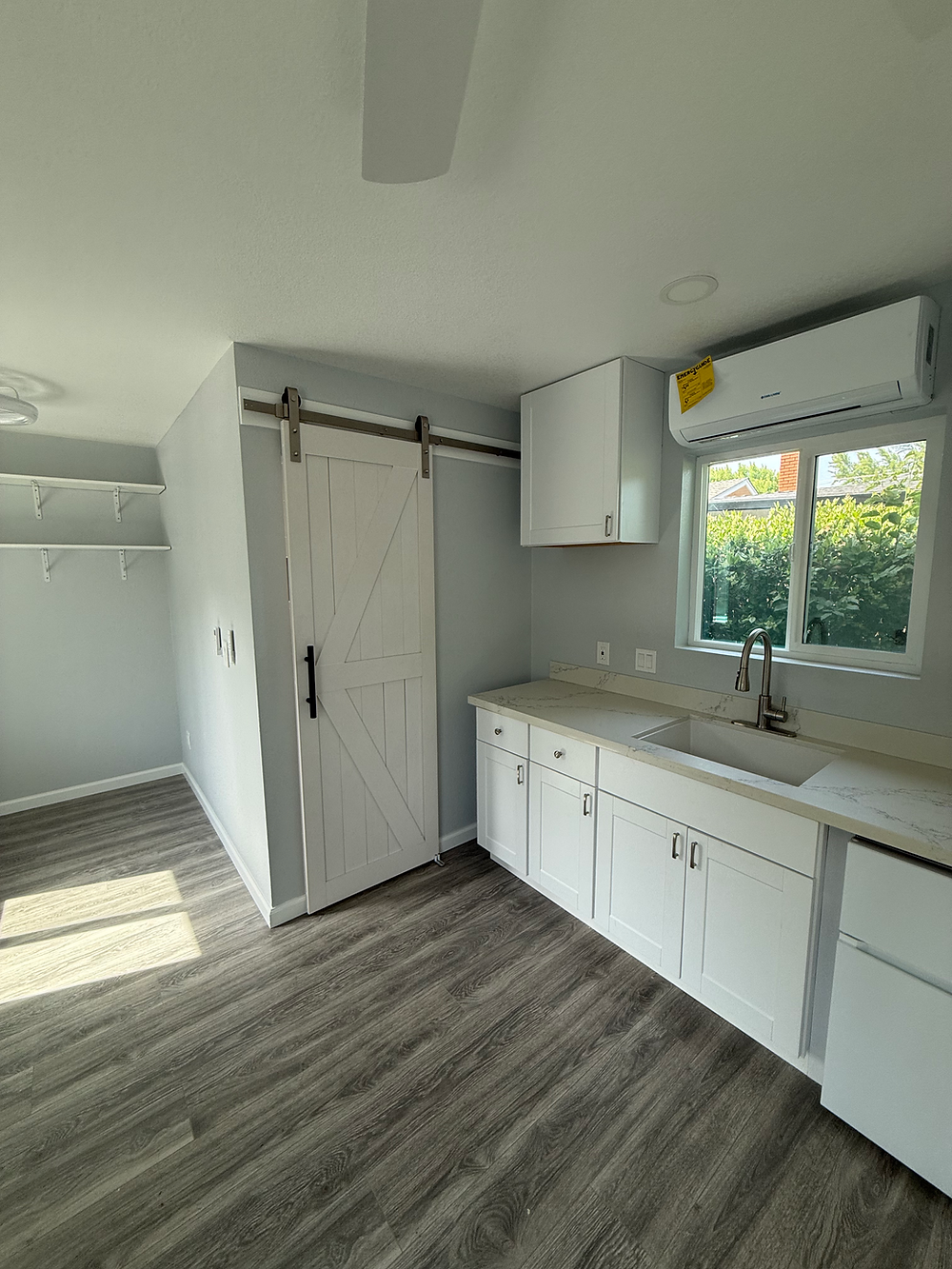 Bright kitchenette remodel with white shaker cabinets, light countertops, gray wood-look flooring, open shelves, and a white sliding barn door.