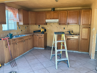 Before kitchen remodel with oak cabinets, tiled floor, missing appliances, patched walls, and tools set out during demolition and renovation work.