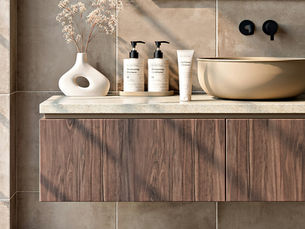 Modern bathroom vanity with floating wood cabinets, a stone countertop, beige tile wall, black wall-mounted faucet, cream vessel sink, and a white vase in warm sunlight.