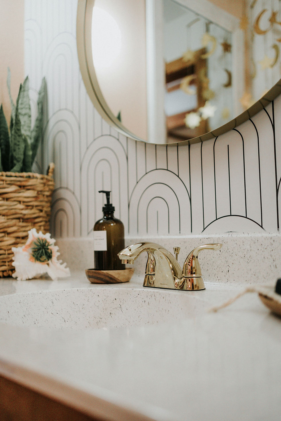 Bathroom sink area with a gold faucet, round mirror, white countertop, patterned arch wallpaper, soap dispenser, woven basket, plant, and small shell decor.