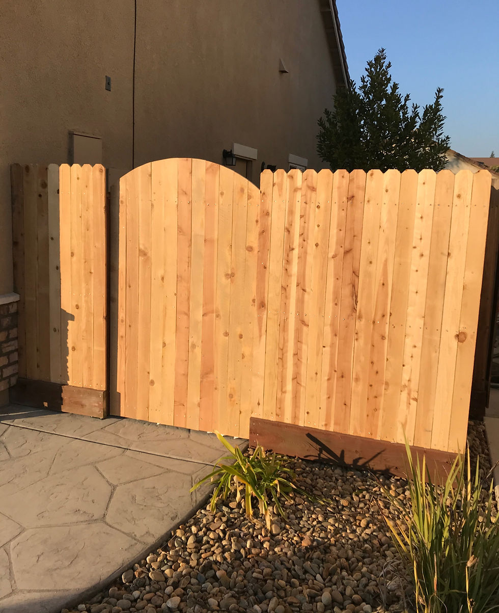 New natural wood fence and gate installed beside a stucco home, with stone landscaping, plants, and a paved walkway in front.