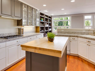 Spacious kitchen with white and gray cabinets, glass-front upper cabinets, granite countertops, wood flooring, large windows, and a butcher block island with a small plant centerpiece.