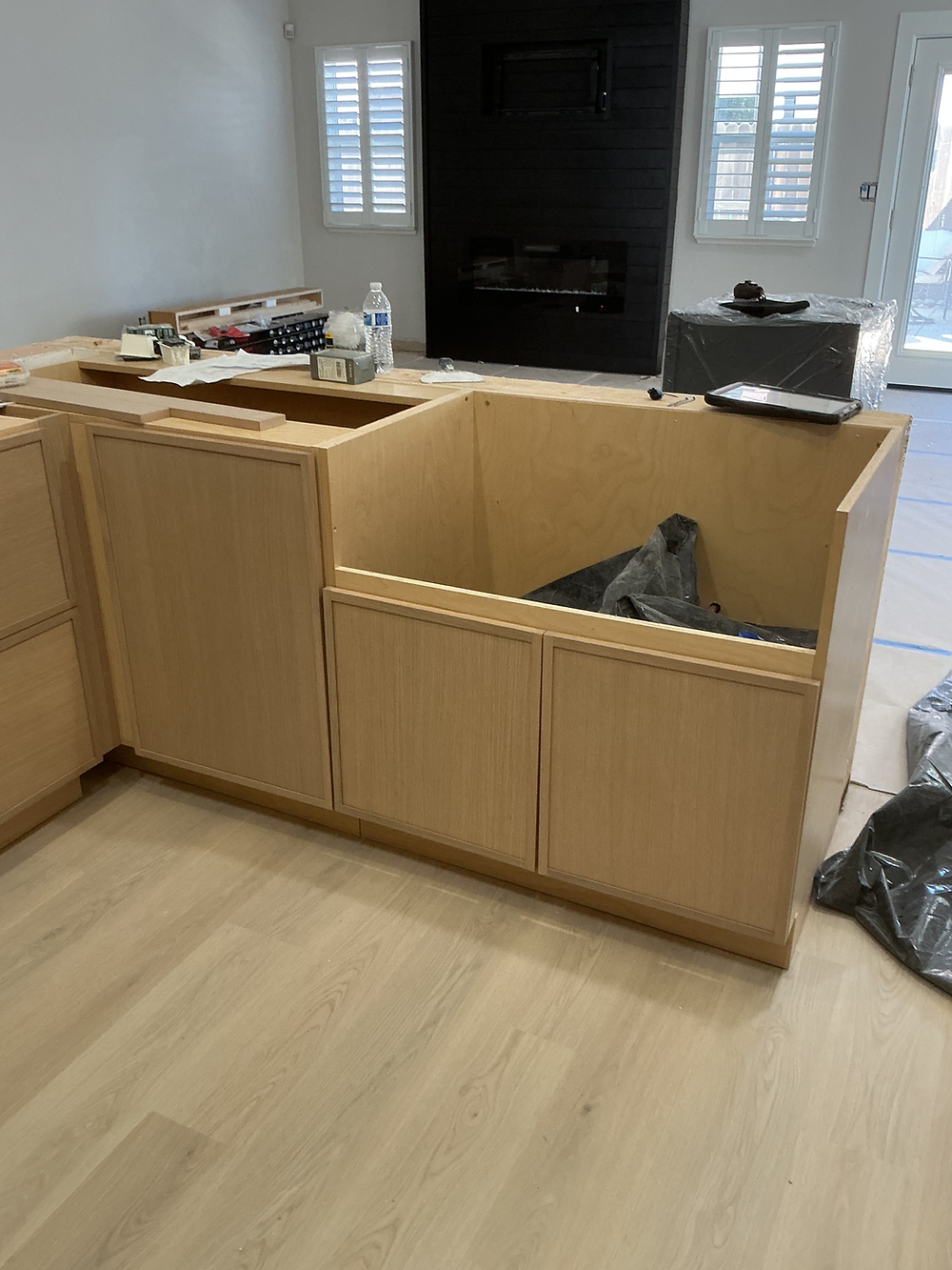 Kitchen island installation in progress with light wood cabinet panels, open countertop framing, updated flooring, and black fireplace wall in the background.