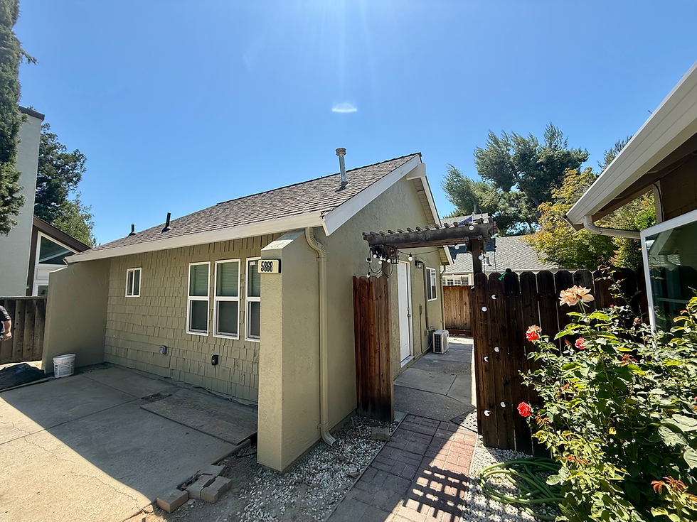 Single-story ADU with light beige siding, white-trimmed windows, and a gabled roof. A brick walkway leads through a wooden pergola to the side entrance, with rose bushes in the foreground.
