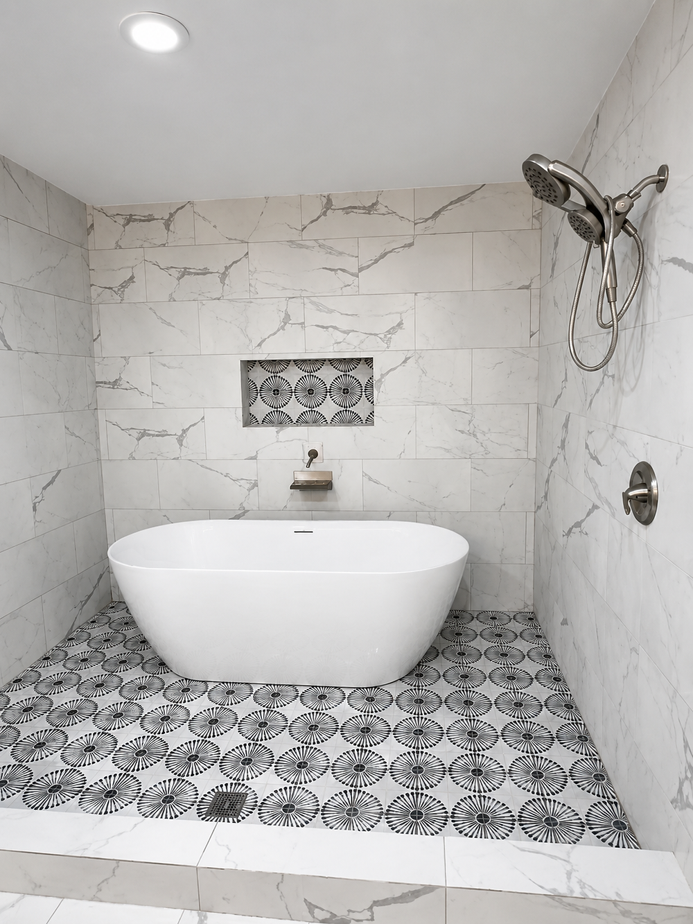 Spa-style bathroom remodel with a freestanding white soaking tub inside a walk-in shower area, marble-look wall tile, patterned floor tile, chrome shower fixture, and recessed wall niche.