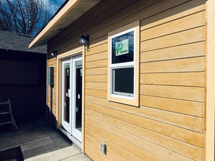 ADU exterior with horizontal wood siding, white-trim sliding glass door, white-trim window, black exterior lights, concrete walkway, and water spigot under a wide roof overhang.