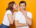 Two coworkers in white t-shirts sharing a secret and reacting playfully—representing team building, workplace communication,
