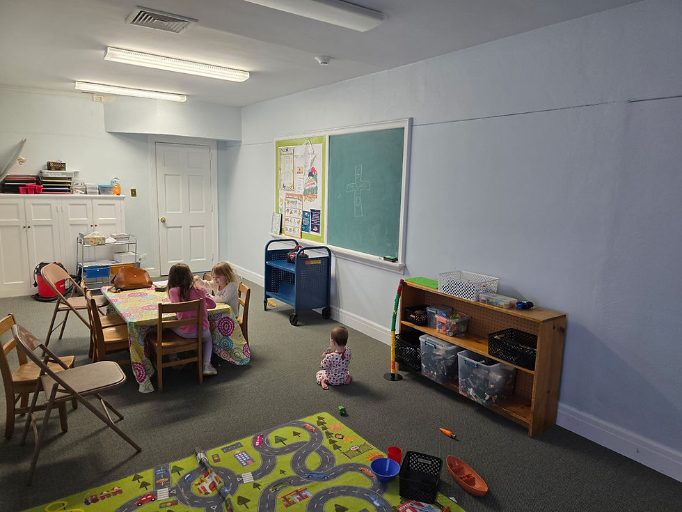 Children in a playroom; two at a table drawing, one on floor near toy shelves. Brightly colored rug, chalkboard with drawings. Calm setting.