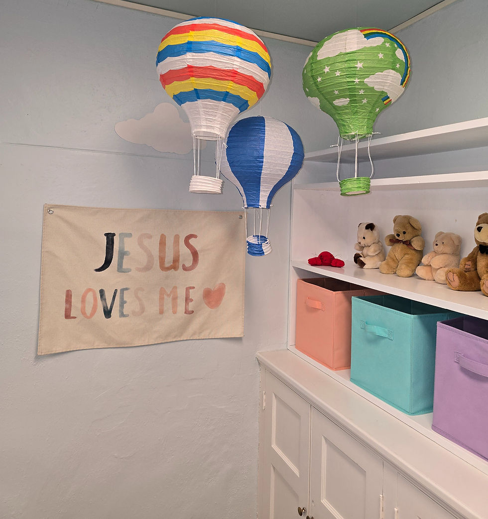 Three colorful paper hot air balloons hang in a room with a "JESUS LOVES ME" sign, pastel storage boxes, and teddy bears on shelves.