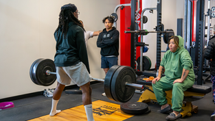 A lifter performs a heavy deadlift on a platform while coaches and teammates observe, illustrating progressive overload through increased weight, focused coaching, and proper strength training form.