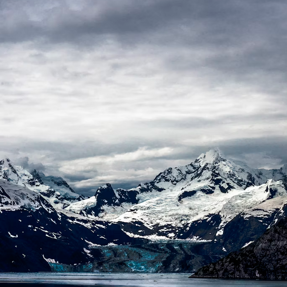 Snow-capped mountains with a glacier overlook a calm, reflective lake under a cloudy, gray sky, creating a serene and cold atmosphere. Part of the Wild Dirt Field Guide National Park Travel Guide Features