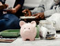 A pink piggy bank and a glass jar filled with cash sit in sharp focus, while two people discuss finances in the blurred background.
