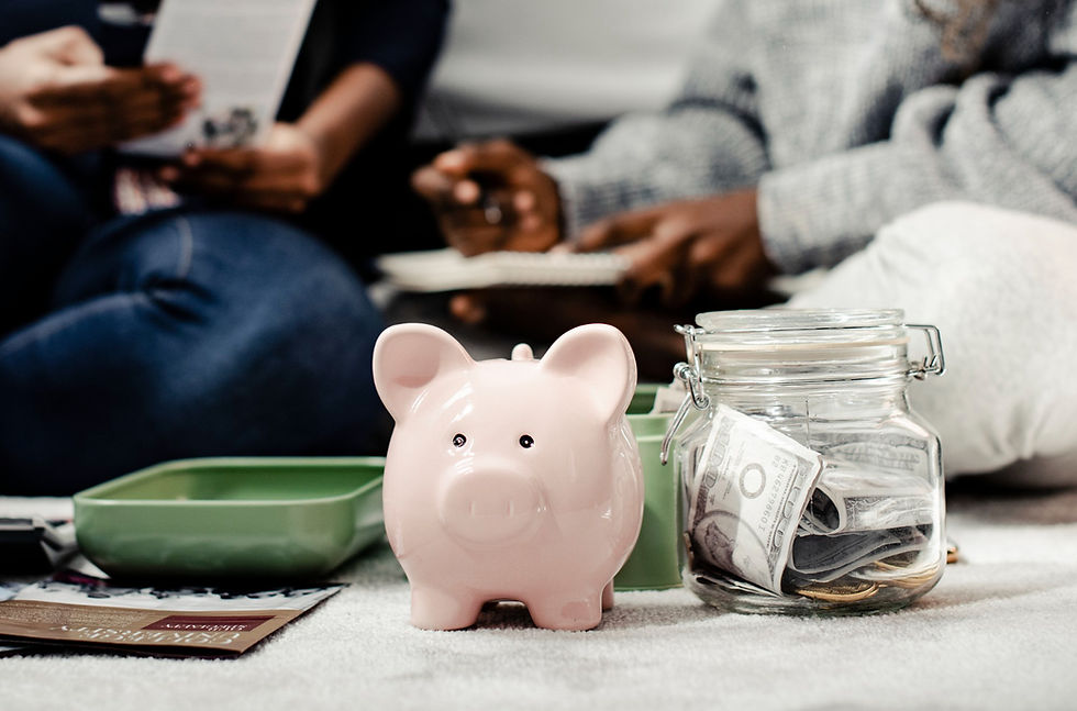 A pink piggy bank and a glass jar filled with cash sit in sharp focus, while two people discuss finances in the blurred background.