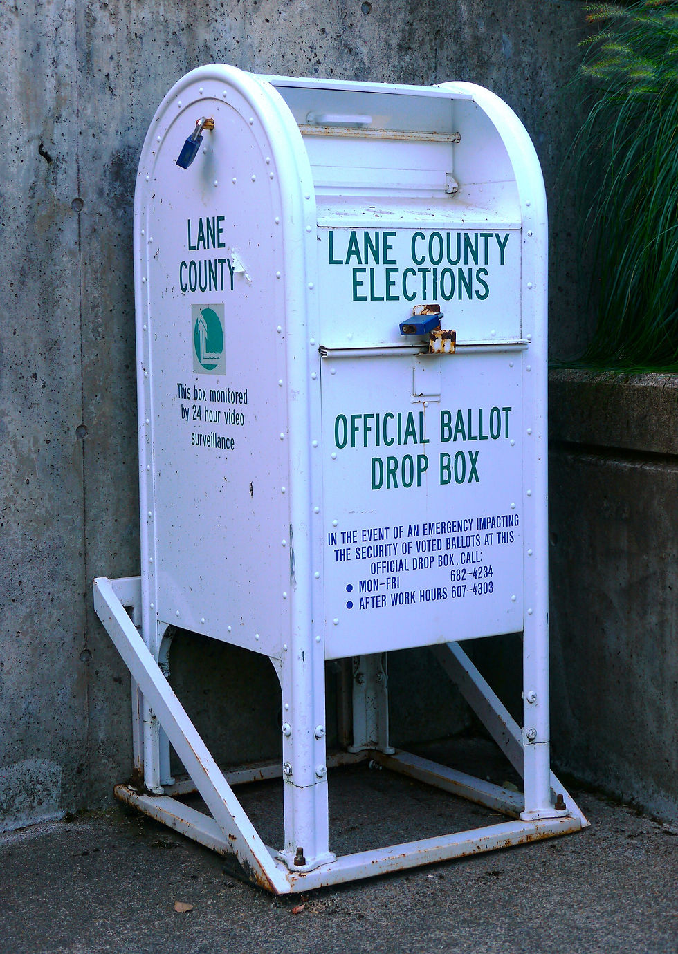 White Lane County Elections official ballot drop box on a metal stand beside a concrete wall