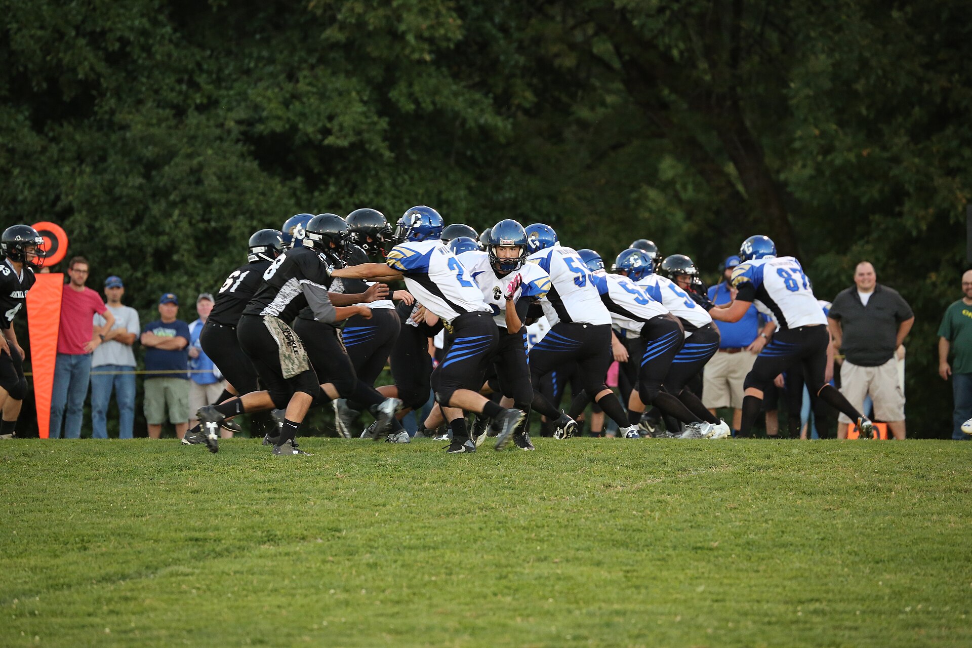 High school football players in black and white uniforms crouch at the line of scrimmage on a grass field as fans watch from the sideline