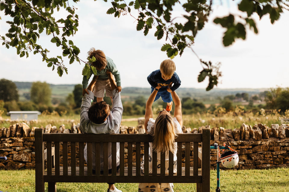 Natural family portrait in the Cotswolds by Felicity Fox, showing parents lifting their children in the air while sitting on a bench outdoors.