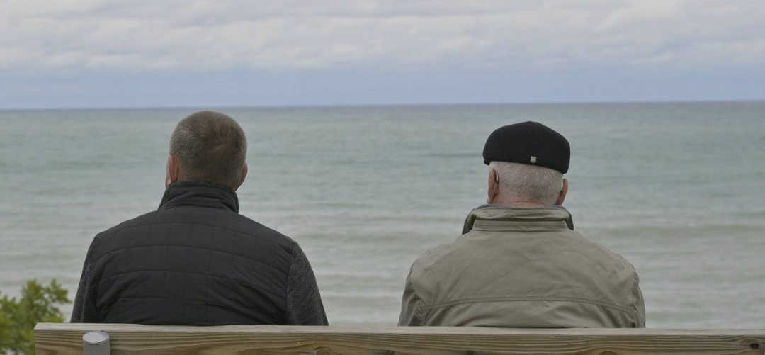 Two men sitting on a bench overlooking the water, representing reflection and connection.