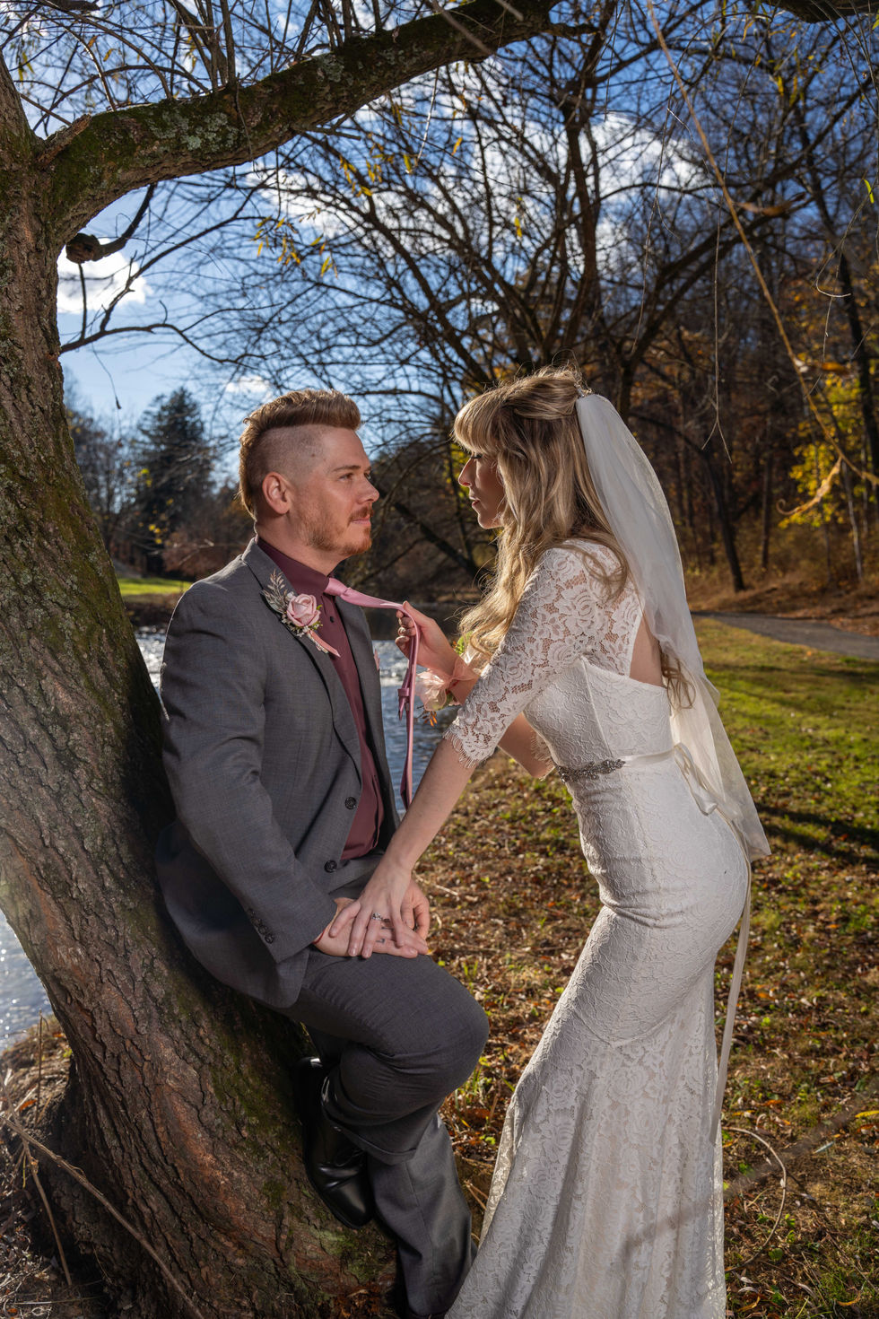 Bride pulling groom's necktie while he leans on tree near river
