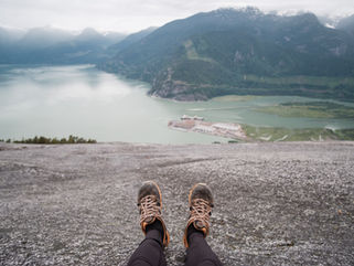 a person's feet in the frame as they look out at a lake from the top of a mountain