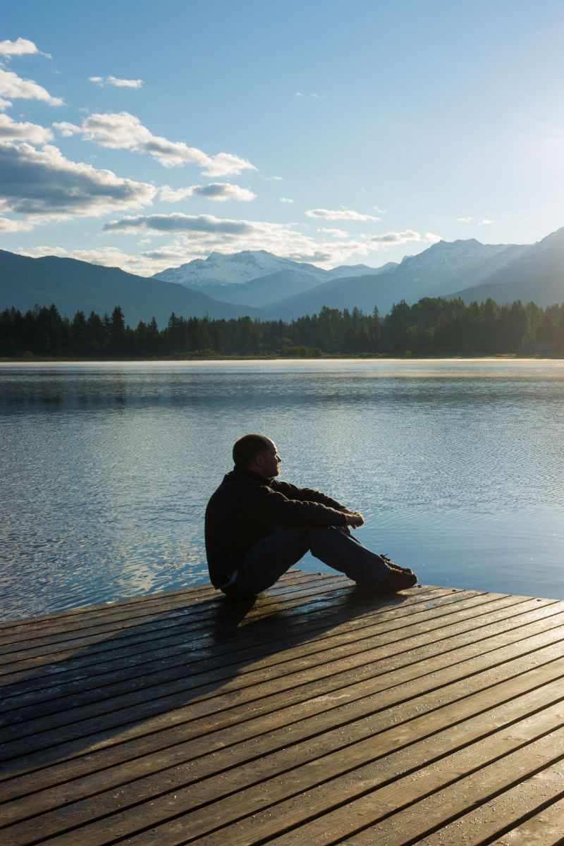 man reflecting on a dock by a smooth lake with mountains in the background