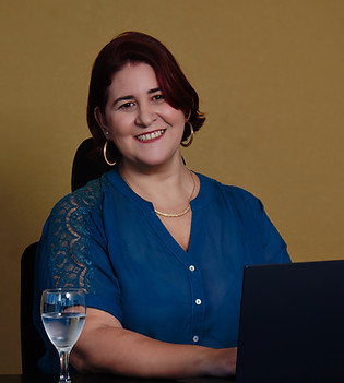 Psicólogoa sentada em frente ao computador, com uma taça de água, sorrindo, olhando para câmera, com uma blusa azul, brincos dourados de argola.