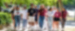 A diverse group of people walk joyfully along a tree-lined path, wearing casual clothes and tees with logos. Sunny day with greenery around.