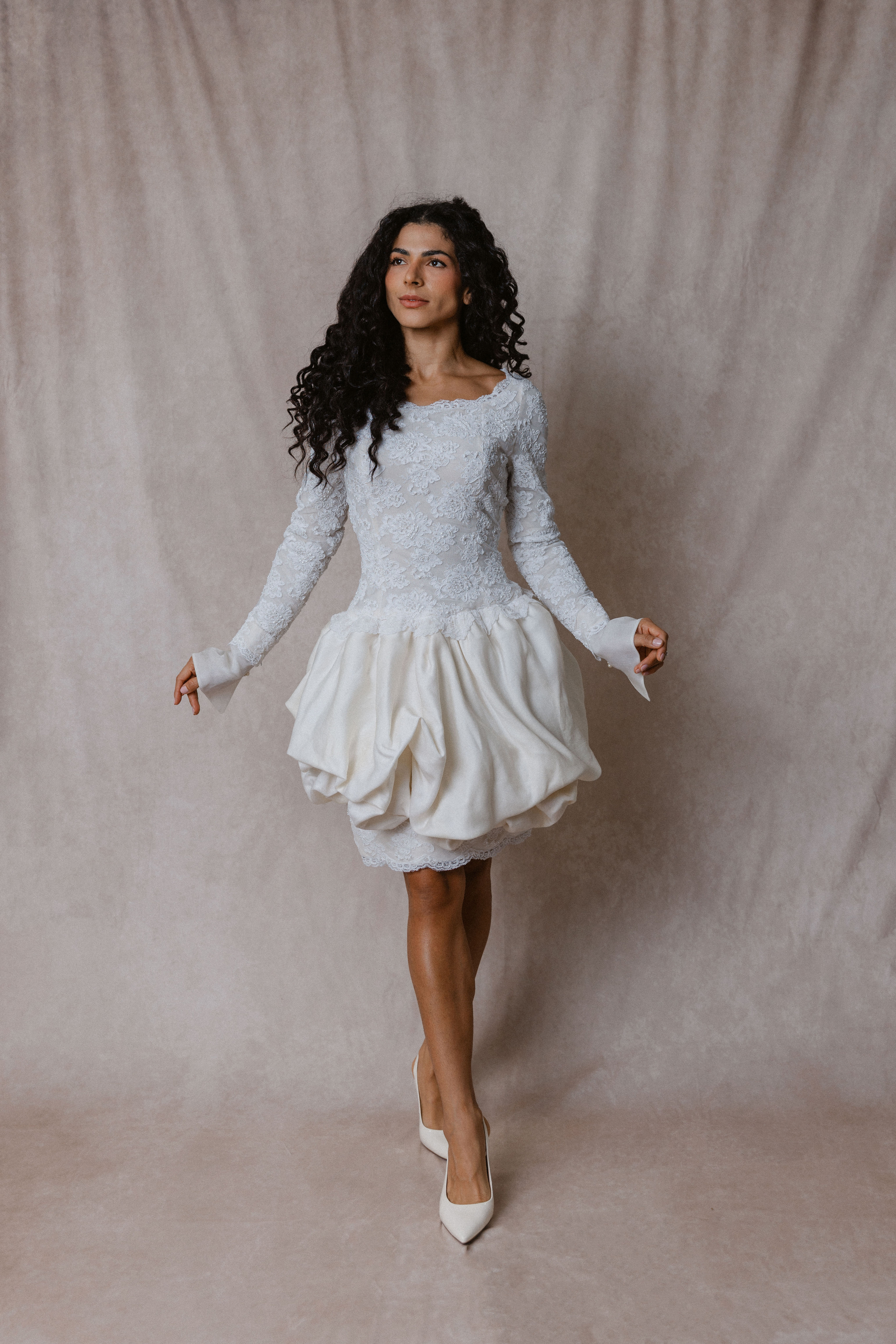 Curly haired model wearing vintage Christian Dior mini wedding dress with lace bodice in front of a beige backdrop