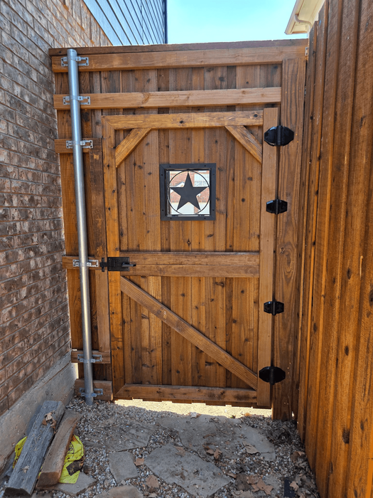 Interior custom wood pedestrian gate with medium-stained vertical boards, diagonal bracing, circular star window, black metal hinges and slide latch, installed beside a brick home.