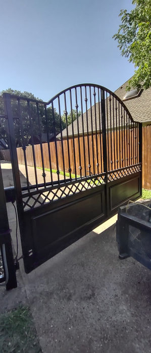Black wrought iron driveway gate with arched top and decorative accents beside a wooden privacy fence