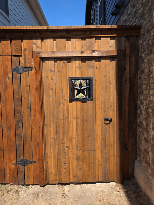 Custom wood pedestrian gate with medium-stained vertical pickets, top cap, square viewing cutout with decorative metal star, and black metal hardware between two residential homes.