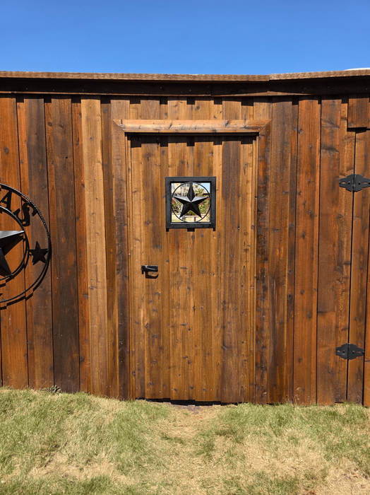 Wood privacy fence with custom pedestrian gate, horizontal top cap, decorative star insert, black metal hardware, and circular metal ornament beside a grass lawn.