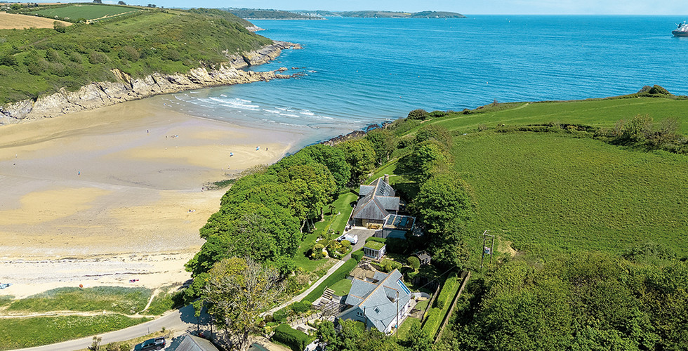 Avalon, a luxury coastal home above Maenporth Beach near Falmouth, Cornwall, offering panoramic sea views across Falmouth Bay. This three storey detached house features a front terrace overlooking the coast, landscaped gardens and a patio designed for al fresco dining. Interior images show a spacious open plan kitchen and dining room with Shaker style cabinetry and freestanding cooker, a glazed conservatory opening onto the garden, a comfortable sitting room with French doors, and a snug with garden views. Four double bedrooms include en suite bathrooms and a dual aspect principal suite, making this an ideal family home in Maenporth. The property also includes an integral garage, utility room, summer house suitable for a home office or studio, and private parking, presented by Jackson-Stops Cornwall with a guide price of £1.3 million.