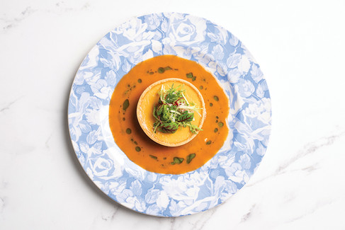 Blue floral plate with a tart surrounded by orange sauce, topped with greens. Set on a white marble surface.